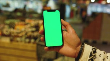 Eller, arka planda bulanık ürünler içeren süpermarket raflarında dikey yeşil ekranlı cep telefonu kullanır. Model dinleme görüntülü kromakey telefon. Yavaş çekim