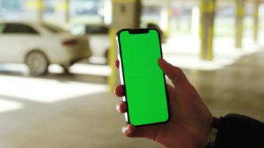 Güneş ışığı parkında, adam telefonu dikey yeşil ekran arka planda beyaz arabalarla tutar. Şehir sokağı akıllı telefon dokunmatik ekranı. Yavaş çekim