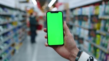 Genç adam cep telefonu kullanıyor. Arka planda bulanık süpermarkette dikey yeşil ekran var. Model dinleme görüntülü kromakey telefon. Yavaş çekim