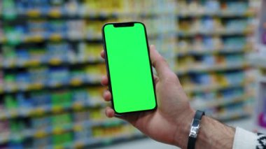 Süpermarketin arka planında dikey yeşil ekranlı cep telefonu kullanan bir adamın el çekimi. Model dinleme görüntülü kromakey telefon. Yavaş çekim