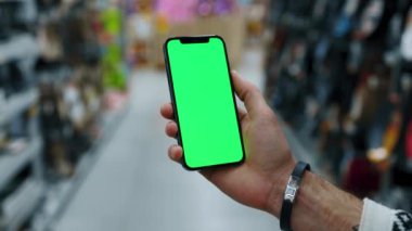 Süpermarkette arka plandaki bulanık adam el kamerasıyla dikey yeşil ekranlı cep telefonu kullanır. Model dinleme görüntülü kromakey telefon. Yavaş çekim