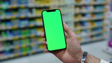 Yavaş çekim, arka planda bulanık süpermarkette dikey yeşil ekranlı cep telefonu kullanır. Model dinleme görüntülü kromakey telefon. Kapat.