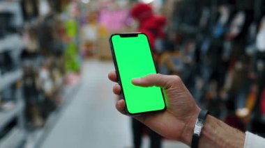 Süpermarketteki bulanık arka plan insanlarında dikey yeşil ekranlı cep telefonu kullan. Model dinleme görüntülü kromakey telefon. Yavaş çekim