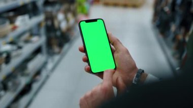 El ele tutuşmada, arka planda bulanık süpermarkette dikey yeşil ekranlı cep telefonu kullan. Model dinleme görüntülü kromakey telefon. Yavaş çekim