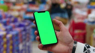 Genç eller, arka planda bulanık süpermarkette dikey yeşil ekranlı cep telefonu kullanır. Kapalı alanda dokunmatik ekranlı müşteri boş cep telefonu. Yavaş çekim
