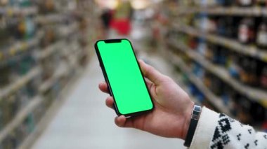 Genç eller akıllı telefon kullanır. Süpermarkette dikey yeşil ekran kullanırlar. Arkaplanı bulanık ve rafları ürünlerle doludur. Model dinleme görüntülü kromakey telefon. Yavaş çekim