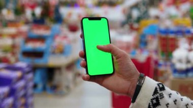 Süpermarkette insan, dikey yeşil ekranlı arka planda ürünlerle bulanık arka plan rafları olan telefonu kullanır. Kapalı alanda dokunmatik ekranlı müşteri boş cep telefonu. Yavaş çekim