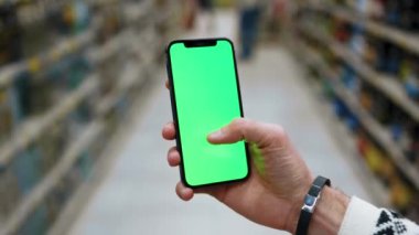 Alışveriş zamanı. Süpermarkette insan, dikey yeşil ekranlı arka planda ürünlerle bulanık arka plan rafları olan telefonu kullanır. Kapalı alanda dokunmatik ekranlı müşteri boş cep telefonu. Yavaş çekim