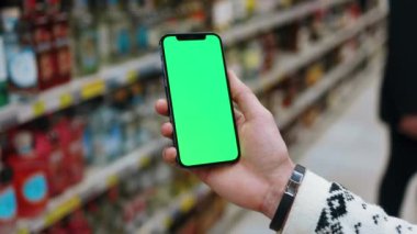 Süpermarkette erkek, arka planı bulanık, dikey yeşil ekranlı telefonu kullanır. Kapalı alanda dokunmatik ekranlı müşteri boş cep telefonu. Yavaş çekim