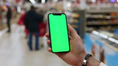 Kapalı mekanlarda erkekler arka planda bulanık insanlar yürürken dikey yeşil ekranlı akıllı telefon kullanırlar. Model dinleme görüntülü kromakey telefon. Yavaş çekim