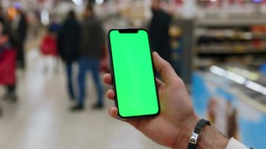 Yavaş çekim yapan adam arka planda bulanık insanlar yürürken dikey yeşil ekranlı akıllı telefonu kullanır. Model dinleme görüntülü kromakey telefon. Kapat.