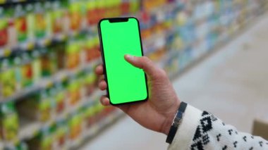 Süpermarkette insan, dikey yeşil ekranlı arka planda ürünlerle bulanık arka plan rafları olan telefonu kullanır. Kapalı alanda dokunmatik ekranlı müşteri boş cep telefonu. Alışveriş zamanı. Kapatın. Yavaş çekim