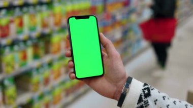 Tezgahtarda, dikey yeşil ekranlı arka planda ürünlerle bulanık arka plan rafları olan telefonu kullanın. Kapalı alanda dokunmatik ekranlı müşteri boş cep telefonu. Alışveriş zamanı. Kapatın. Yavaş çekim