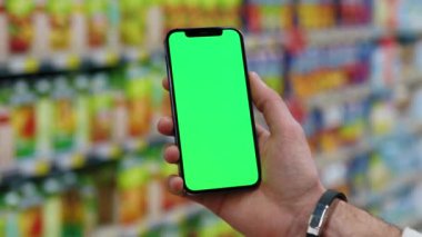 Yavaş çekim adamı, dikey yeşil ekranlı arka planda ürünlerle bulanık arka plan rafları olan telefonu kullanır. Kapalı alanda dokunmatik ekranlı müşteri boş cep telefonu. Alışveriş zamanı. Kapat.