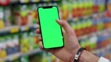Adam elleri ile adam dikey yeşil ekranlı arka planda ürünlerle bulanık arka plan rafları kullanarak telefonu tutar. Kapalı alanda dokunmatik ekranlı müşteri boş cep telefonu. Alışveriş zamanı. Kapatın. Yavaş çekim