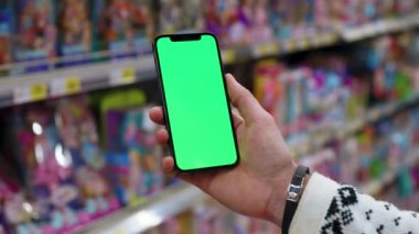 Mağazada erkek, dikey yeşil ekranlı arka planda ürünlerle bulanık arka plan rafları olan telefonu kullanır. Kapalı alanda dokunmatik ekranlı müşteri boş cep telefonu. Alışveriş zamanı. Kapatın. Yavaş çekim