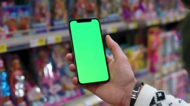 Elle çekilen adam, dikey yeşil ekranlı arka planda ürünlerle bulanık arka plan rafları kullanarak telefonu tutar. Kapalı alanda dokunmatik ekranlı müşteri boş cep telefonu. Alışveriş zamanı. Dükkanda. Yavaş çekim