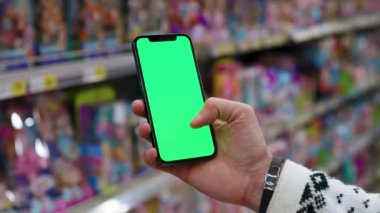 Eller tutucu, dikey yeşil ekranlı arka planda ürünlerle bulanık arka plan rafları olan telefonu kullanır. Kapalı alanda dokunmatik ekranlı müşteri boş cep telefonu. Alışveriş zamanı. Dükkanda. Yavaş çekim