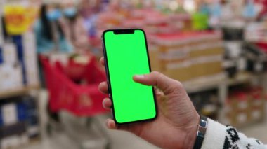 Alışveriş merkezinde çalışan bir adam, dikey yeşil ekranlı arka planda ürünlerle bulanık arka plan rafları olan telefonu kullanır. Kapalı alanda dokunmatik ekranlı müşteri boş cep telefonu. Alışveriş zamanı. Kapatın. Yavaş çekim