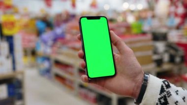 Eller tutucu, dikey yeşil ekranlı arka planda ürünlerle bulanık arka plan rafları olan telefonu kullanır. Kapalı alanda dokunmatik ekranlı müşteri boş cep telefonu. Alışveriş zamanı. Kapatın. Yavaş çekim