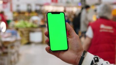 Eller tutucu, dikey yeşil ekranlı arka planda insanların yürüdüğü ürünlerle bulanık arka plan rafları olan telefonu kullanır. Kapalı alanda dokunmatik ekranlı müşteri boş cep telefonu. Alışveriş zamanı. Kapatın. Yavaş çekim
