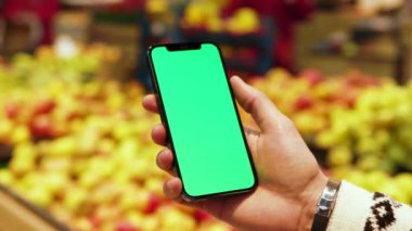Erkek eli, arka planda bulanık meyveler içeren dikey yeşil ekranlı cep telefonu kullanır. Model dinleme görüntülü kromakey telefon. Dükkanda. Kapatın. Yavaş çekim