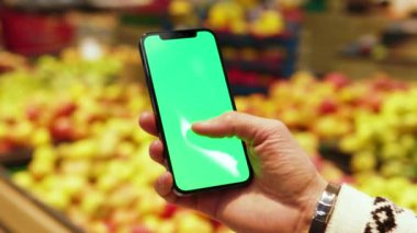 Eller, arka planda bulanık meyveler içeren, dikey yeşil ekranlı cep telefonu kullansın. Model dinleme görüntülü kromakey telefon. Dükkanda. Kapatın. Yavaş çekim