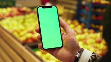Süpermarkette cep telefonu kullanırken arka planda bulanık yeşil ekran meyveler kullanılıyor. Model dinleme görüntülü kromakey telefon. Dükkanda. Kapatın. Yavaş çekim