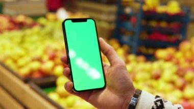 Eller, arka planda bulanık meyveler içeren, dikey yeşil ekranlı cep telefonu kullansın. Model dinleme görüntülü kromakey telefon. Dükkanda. Kapatın. Yavaş çekim