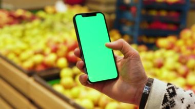 Eller yukarı, arka planda bulanık meyveler içeren dikey yeşil ekranlı cep telefonu kullanın. Model dinleme görüntülü kromakey telefon. Dükkanda. Yavaş çekim