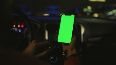 Gece vakti el kamerasıyla çekilen fotoğraf. Arabada dikey yeşil ekranlı akıllı telefon kullanılıyor. Cihaz dijital cihaz, dokunmatik ekranlı mobil telefon teknolojisi. Yavaş çekim