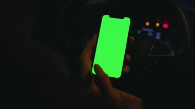 Genç adamın gece vakti arabasında dikey yeşil ekranlı akıllı telefon kullanması. Cihaz dijital cihaz, dokunmatik ekranlı mobil telefon teknolojisi. Yavaş çekim
