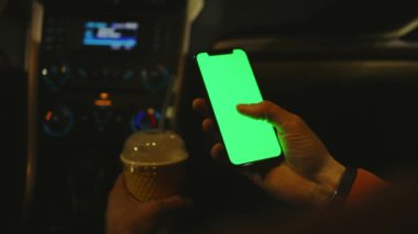 Arabada çalışan bir adam geceleri kahve içmek için dikey yeşil ekranlı akıllı telefon kullanır. Cihaz dijital cihaz, dokunmatik ekranlı mobil telefon teknolojisi. Yavaş çekim