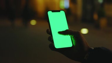 Yavaş çekim yapan adam, gece caddesinde dikey yeşil perdeli telefon kullanıyor. İyi akşamlar akıllı telefon ekran uygulaması. Teknoloji dokunmatik hücre ekranı. Kapat.