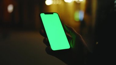 Elle çekilen adam gece şehrinde dikey yeşil ekran standı olan telefonu kullanıyor. İyi akşamlar akıllı telefon ekran uygulaması. Teknoloji dokunmatik hücre ekranı. Yavaş çekim