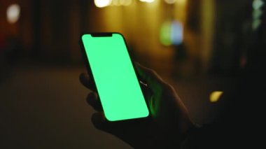 Sokakta çalışan adam, gece şehrinde dikey yeşil ekran standı olan telefonu kullanır. İyi akşamlar akıllı telefon ekran uygulaması. Teknoloji dokunmatik hücre ekranı. Yavaş çekim