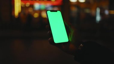 Eller gece şehir arka planında dikey yeşil ekranlı telefon kullanıyor. İyi akşamlar akıllı telefon ekran uygulaması. Teknoloji dokunmatik hücre ekranı. Yavaş çekim