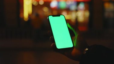 Yavaş çekim yapan adam, gece şehrinde dikey yeşil ekran standı olan telefonu kullanır. İyi akşamlar akıllı telefon ekran uygulaması. Teknoloji dokunmatik hücre ekranı. Kapat.