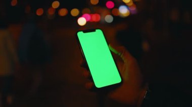 Eller gece şehir arka planında dikey yeşil ekranlı telefon kullanıyor. İyi akşamlar akıllı telefon ekran uygulaması. Teknoloji dokunmatik hücre ekranı. Yavaş çekim