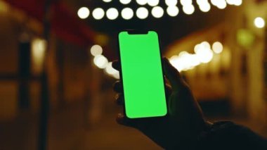 Geceleri arka plandaki çelenklerde dikey yeşil ekranlı cep telefonu tutan bir adam. Mobil internet bağlantısı teknolojisi cihaz hücresi. Yavaş çekim