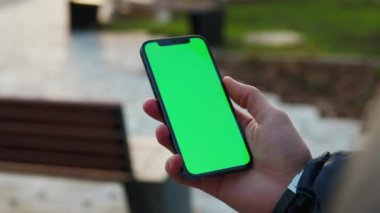 NEW YORK - 19 Aralık 2020: Gün ışığında, arka plandaki bankta dikey yeşil ekranlı telefonu tutan genç adam. İnternet parmak aleti. Yavaş çekim