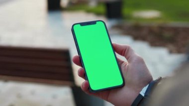 NEW YORK - 19 Aralık 2020: Park arka planındaki bankta dikey yeşil ekranlı telefonu tutan genç elleri kapatın. İnternet parmak aleti. Yavaş çekim