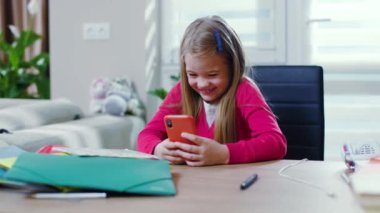 Evde oturup ödev yapan gülümseyen kız telefonu kullan, mutlu ol. Liseli kız portresi karantina altında salgın üzerinde çalışıyor. Masanın yanında oturuyor. Yavaş çekim.