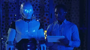 Genç Afro-Amerikan iş adamı portresi. Sibernetik robotlarla konuşuyor. Şeffaf ekran kullanıyor. Geleceğin çalışanları. Robot ve insan işbirliği. İletişim, proje, gelişme. Yavaş