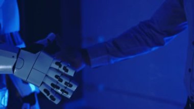 Bir iş adamının mavi ışıklı elleriyle sibernetik bir robotla el sıkışıyor. Yapay zekayla insan etkileşimi. Ortaklık, gelecek, üretkenlik, anlaşma, bilim. Yavaş çekim