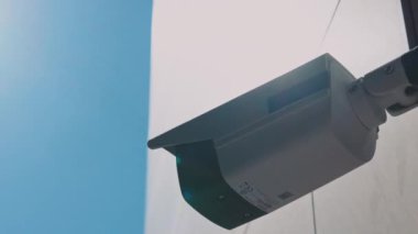 Mavi gökyüzü arka planındaki güvenlik kamerası gün ışığında bir binanın dışında kayıt yapıyor. Güvenlik kameraları. Mahremiyet, şehir, koruma, ev, sistem, kontrol, güvenlik, görüş, güvenlik, video. Yavaş çekim