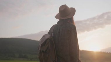 Sırt çantalı, çayırda gün batımını izleyen, şapkalı ve çantalı, sıcak altın çantalı yalnız gezginlerin yavaş sinematik tavası.
