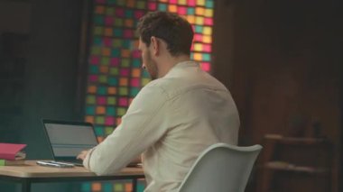 Bilgisayardaki adam kazandıktan sonra başarıyı kutluyor, parlak cam pencereler ve modern ofiste doğal ışık, kağıtlar.