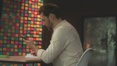 Masada oturan adam not defterine notlar yazıyor ve fikirler çiziyor. Stüdyoda doğal pencere ışığı, kalemler ve kağıtlar var.