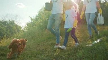 Beyaz aile çevre ekibi çimenli yamaçta fidan dikiyor ve suluyor. Çocuk su kovası taşırken ebeveynler kazıyor.